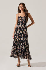 A woman is wearing a sleeveless, black maxi dress with thin spaghetti straps. The dress features a floral pattern with large white and pink flowers scattered across the fabric. The hem of the dress is ruffled, adding a flowing, feminine touch.