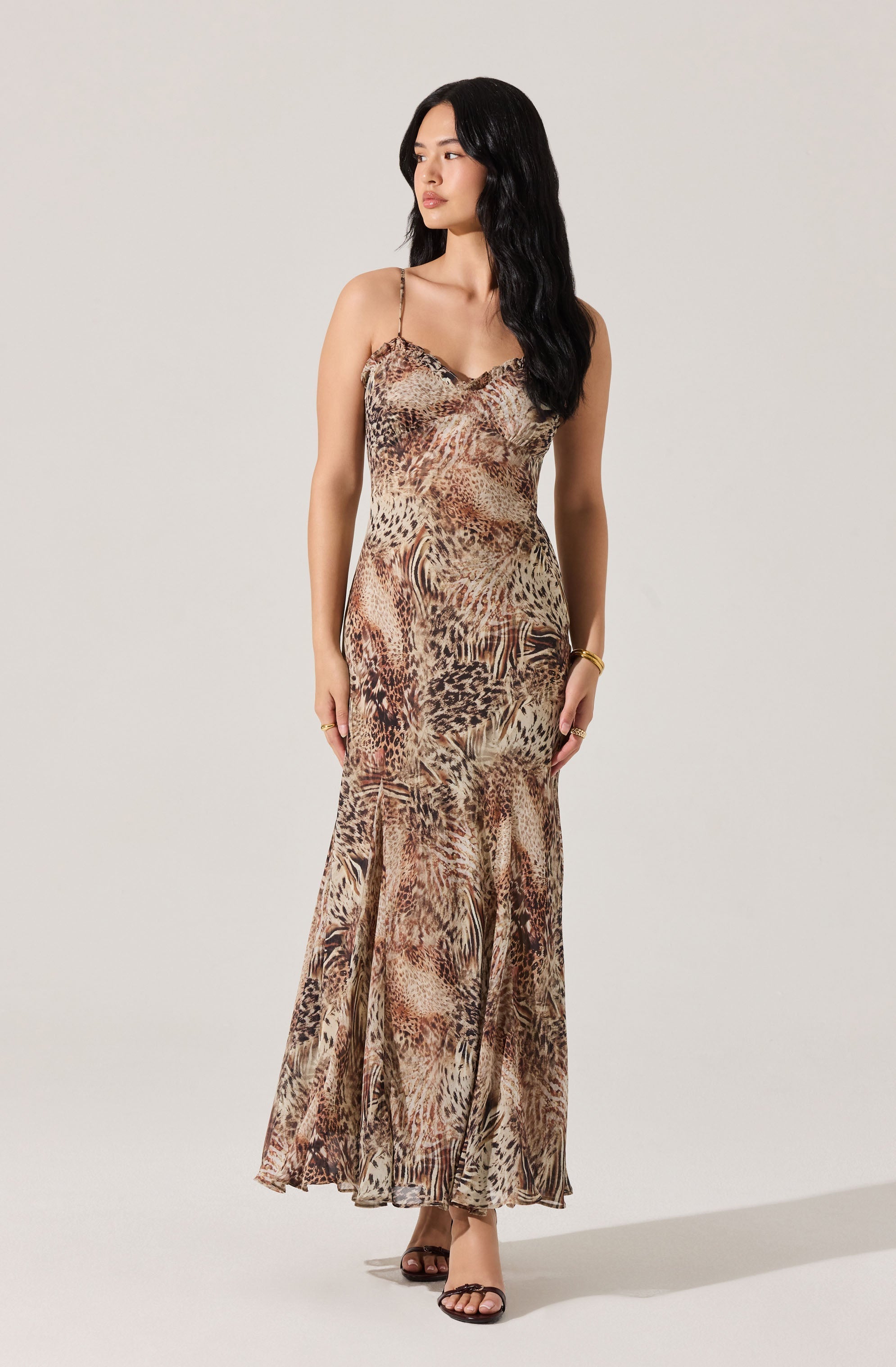 The woman is wearing a sleeveless, full-length dress with a fitted bodice and a flowing skirt. The dress features a multicolored animal print pattern, including shades of brown, beige, and black, resembling various wild animal spots and stripes. The dress has thin spaghetti straps and a slightly ruffled neckline. She is also wearing open-toed high-heel sandals and a bracelet on her left wrist.