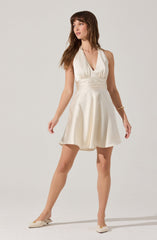 A woman is wearing a sleeveless, V-neck, satin dress in an ivory or champagne color. The dress features a fitted waistband and a flared, A-line skirt. She has paired the dress with matching satin shoes and is accessorized with a gold bracelet.