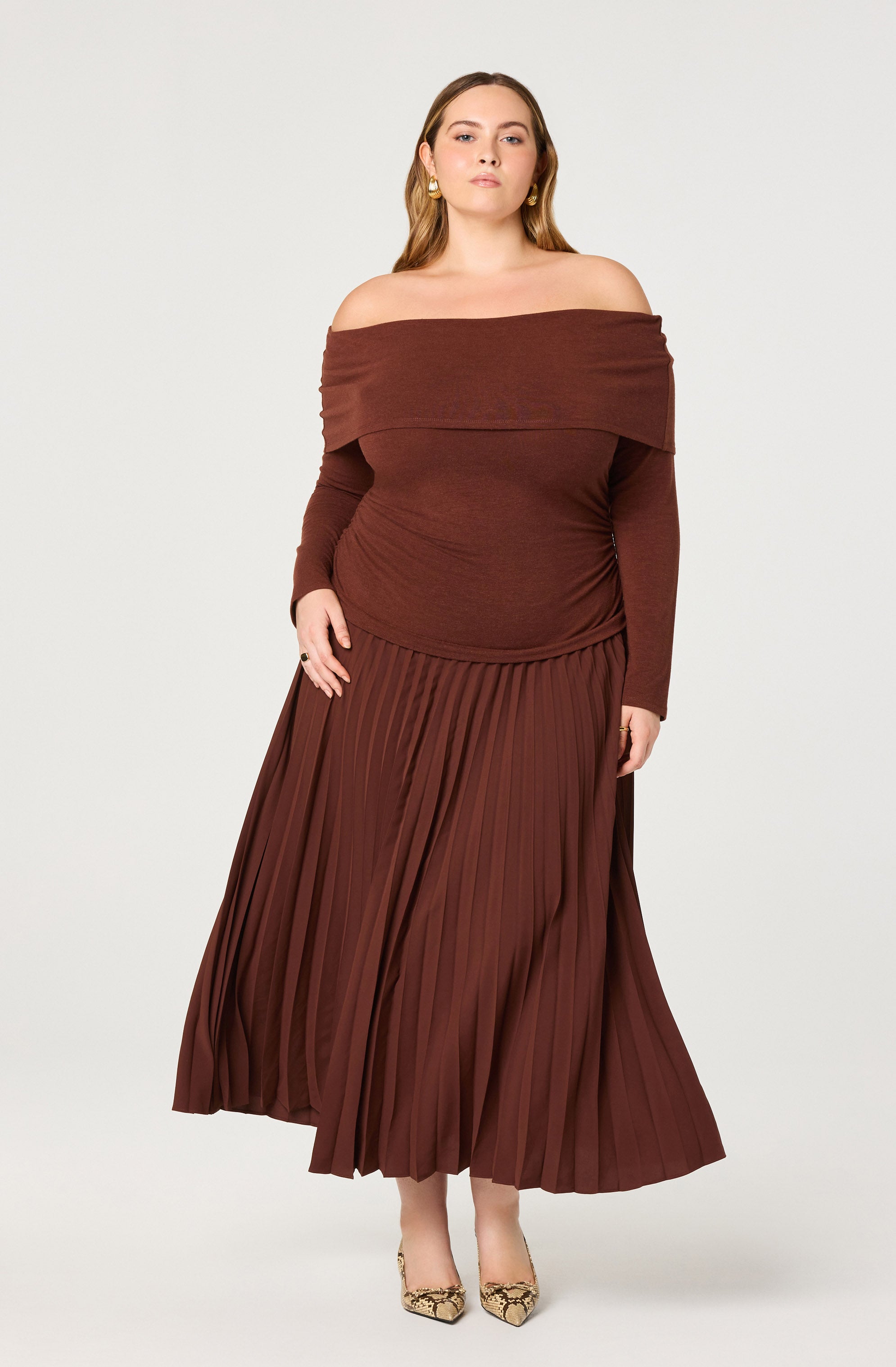A woman is wearing an off-the-shoulder, long-sleeved dress in a deep brown color. The top part of the dress is fitted with a wide, folded neckline that drapes around her shoulders. The skirt features a pleated design that extends to her ankles, creating a flowing silhouette. She is also wearing patterned pointed-toe shoes.