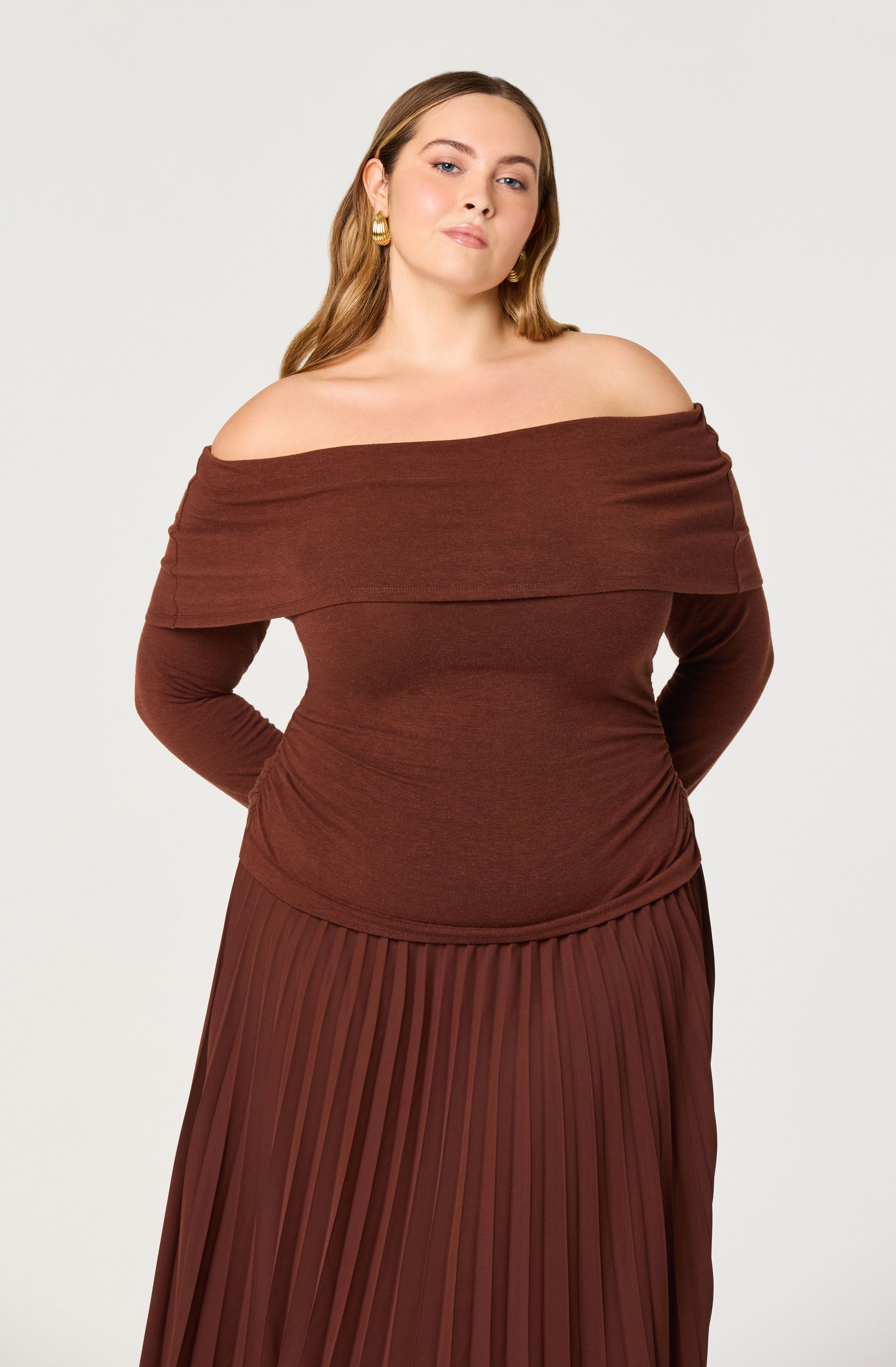 A woman wearing an off-the-shoulder, long-sleeve top in a rich brown color, with a fold-over neckline. She is also wearing a matching pleated skirt in the same brown shade.