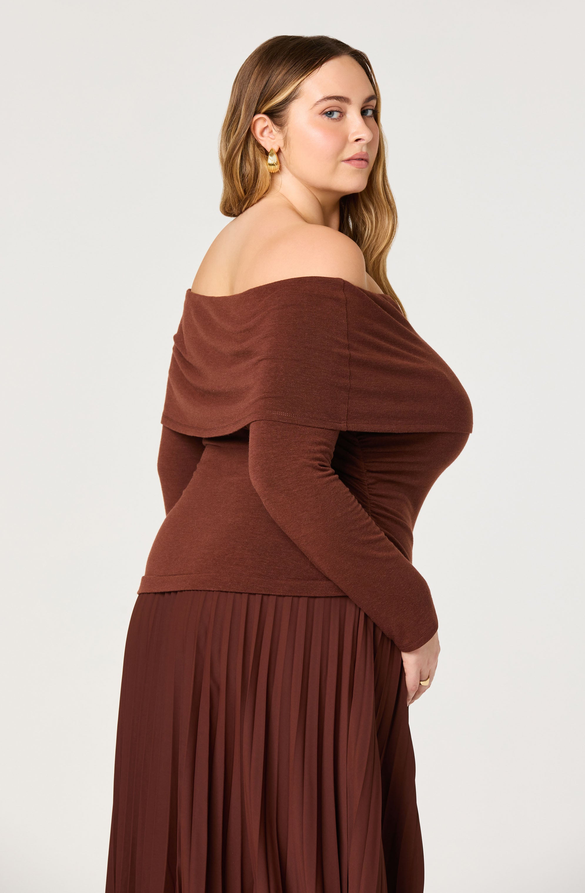 A woman is wearing a brown, off-the-shoulder top with a wide, draped neckline that exposes her shoulders and collarbone. She is also dressed in a matching brown pleated skirt that reaches below her knees. She has gold earrings and her hair is styled in loose waves.
