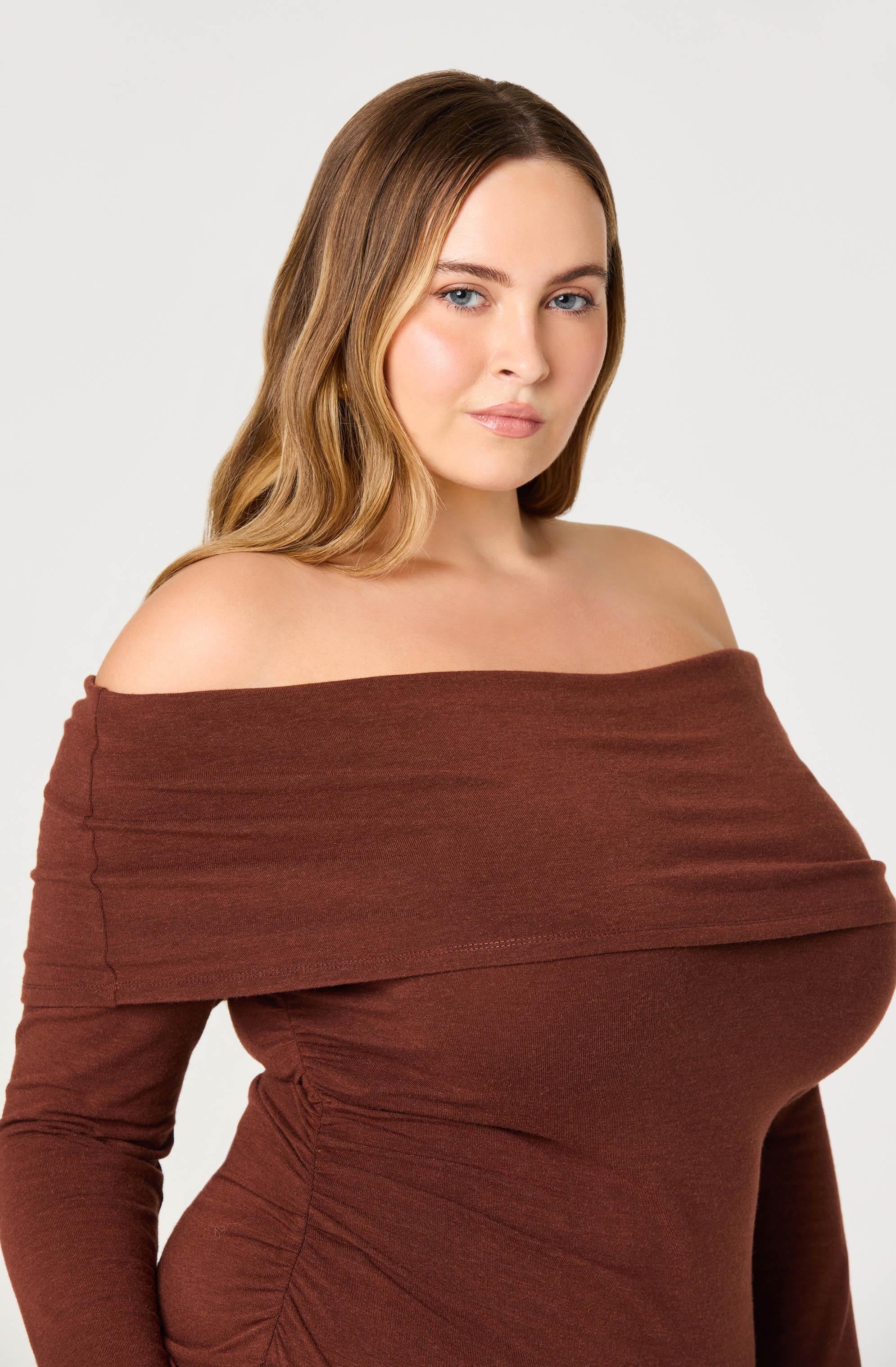 A woman wearing an off-the-shoulder, long-sleeved top in a deep brown color. The top is fitted and features ruching details along the sides.