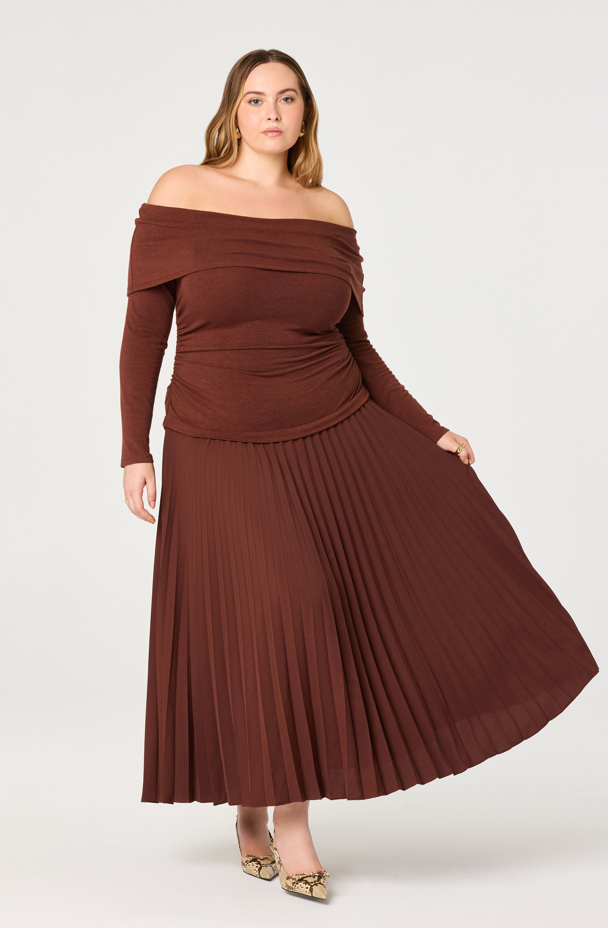 A woman is wearing a matching brown off-the-shoulder top with long sleeves and a wide neckline that drapes over her shoulders. She pairs it with a high-waisted, pleated, floor-length skirt in a similar brown shade, creating a coordinated look. She completes her outfit with patterned high heels.