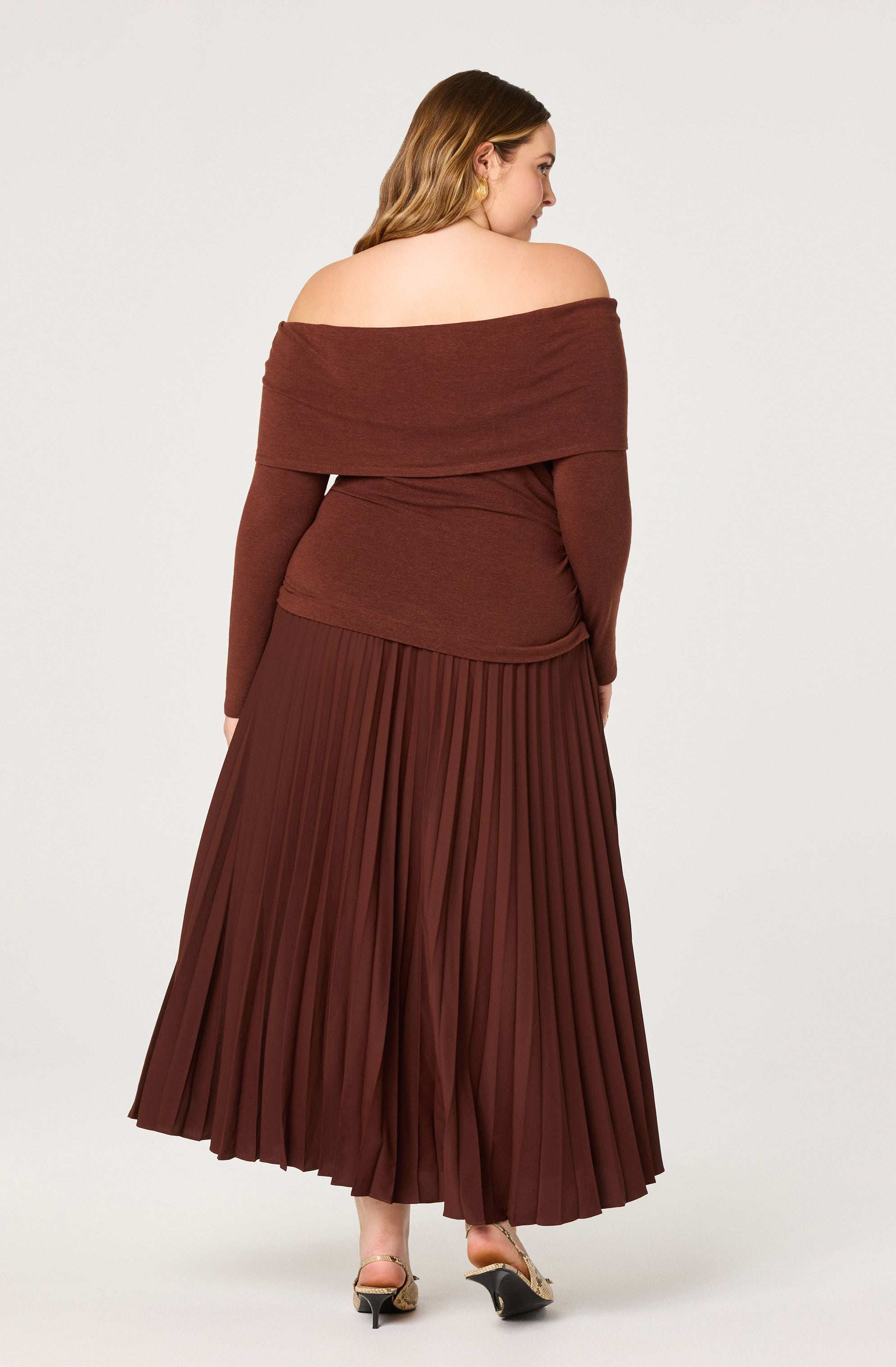 A woman is wearing an off-the-shoulder, long-sleeved top in a warm brown color. The top appears to be made of a soft, knit fabric and has a wide neckline that drapes over her shoulders. She is also wearing a matching maxi skirt with a pleated design in a similar brown shade. The skirt has a flowing, wide-leg silhouette that reaches the floor. She is wearing open-toe, high-heeled shoes with a gold metallic strap.