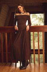 A woman is wearing an off-the-shoulder, long-sleeved, dark brown dress with a fitted bodice and a pleated, floor-length skirt. She accessorizes with a large, chunky silver necklace and a ring on her left hand. She is also wearing black heeled boots.