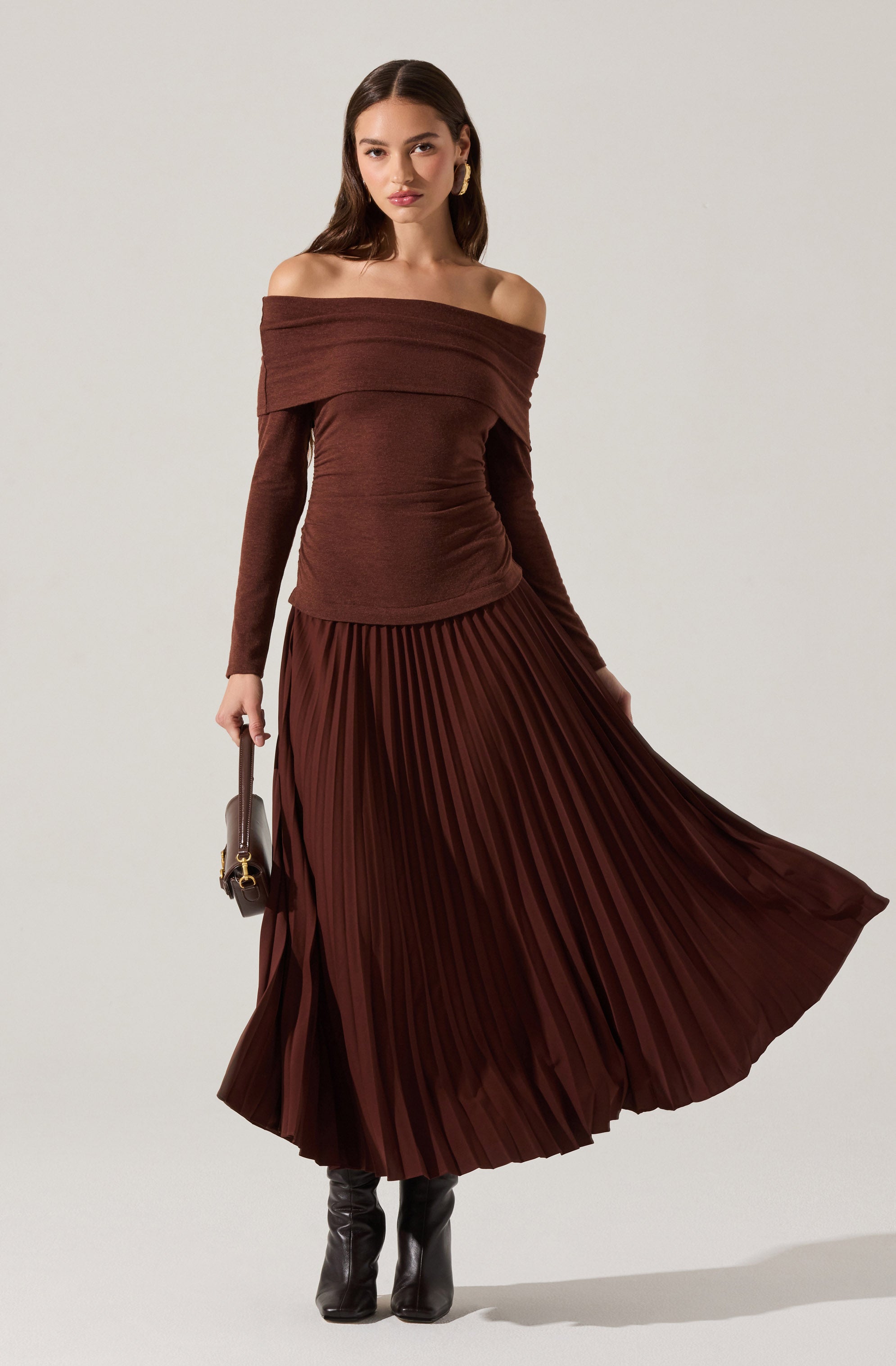 A woman is wearing a matching brown off-the-shoulder sweater and a long, pleated skirt. The sweater has a wide neckline that drapes over her shoulders and long sleeves. The skirt is floor-length with narrow, vertical pleats. She is also wearing black boots and holding a small handbag in her left hand.