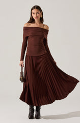 A woman is wearing a matching brown off-the-shoulder sweater and a long, pleated skirt. The sweater has a wide neckline that drapes over her shoulders and long sleeves. The skirt is floor-length with narrow, vertical pleats. She is also wearing black boots and holding a small handbag in her left hand.