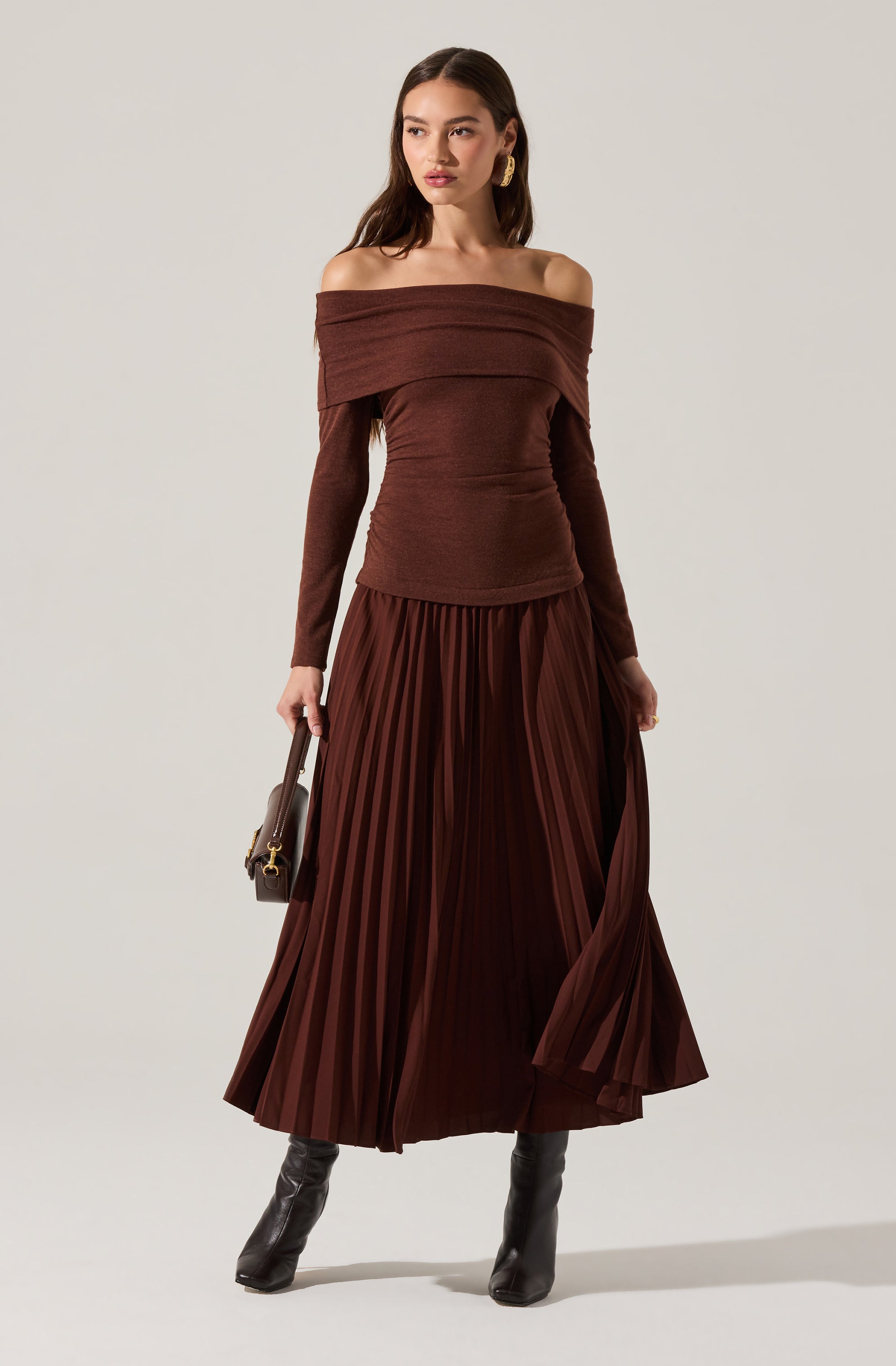 A woman is wearing an off-the-shoulder, long-sleeved brown top with a wide neckline. She pairs it with a matching high-waisted, pleated, floor-length skirt in a rich brown color. She accessorizes with large gold hoop earrings, black knee-high boots, and carries a small dark handbag with a short handle.