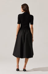 A woman is wearing a fitted, short-sleeved black top made of ribbed fabric, paired with a voluminous, knee-length black skirt that has a gathered waist. She is also wearing black high-heeled shoes.