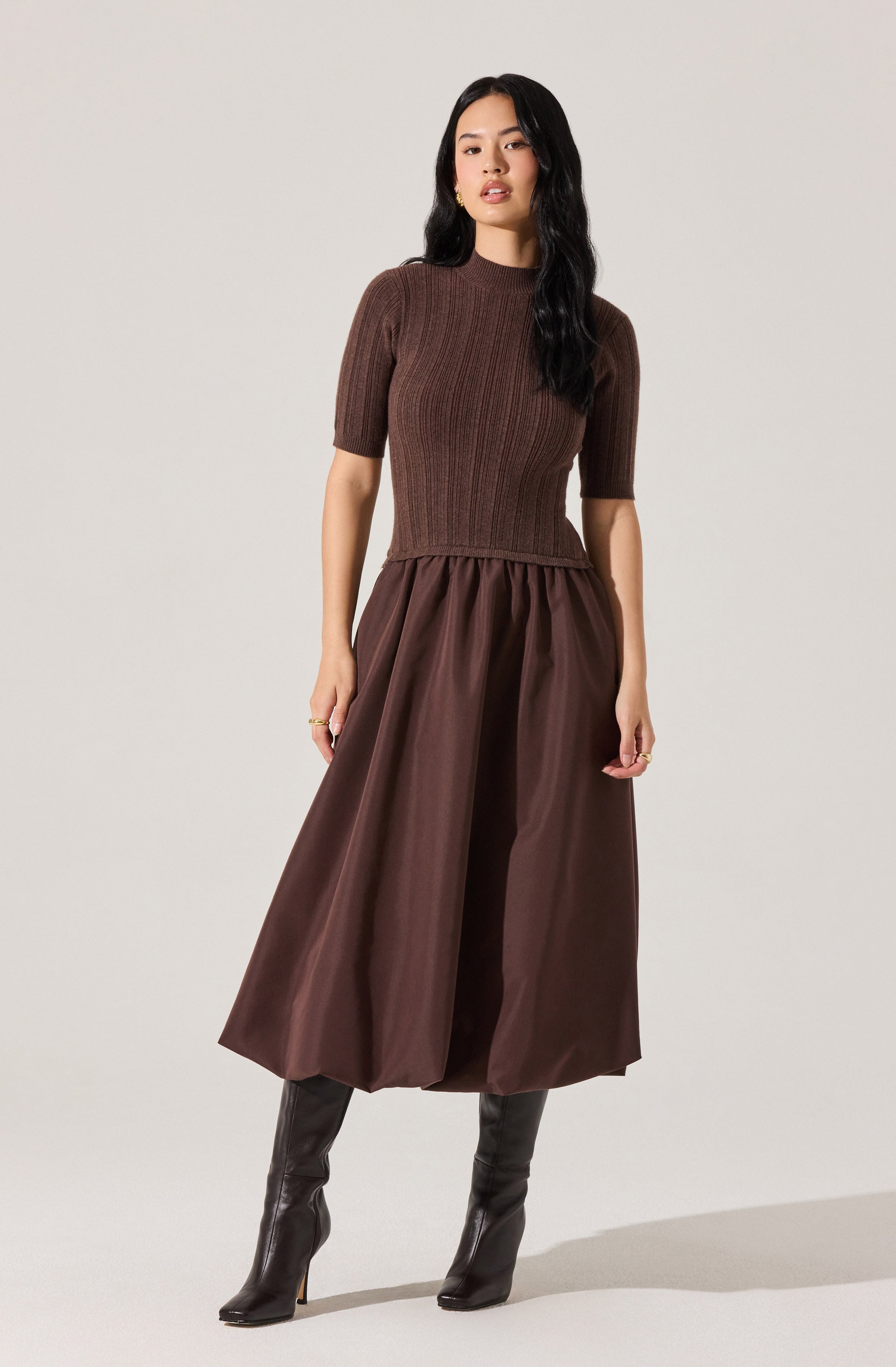A woman is wearing a fitted, short-sleeved, ribbed knit top in a dark brown color with a high neckline. She pairs it with a voluminous, ankle-length skirt in a matching dark brown satin fabric, creating a monochromatic outfit. She completes her look with a pair of tall, black heeled boots.