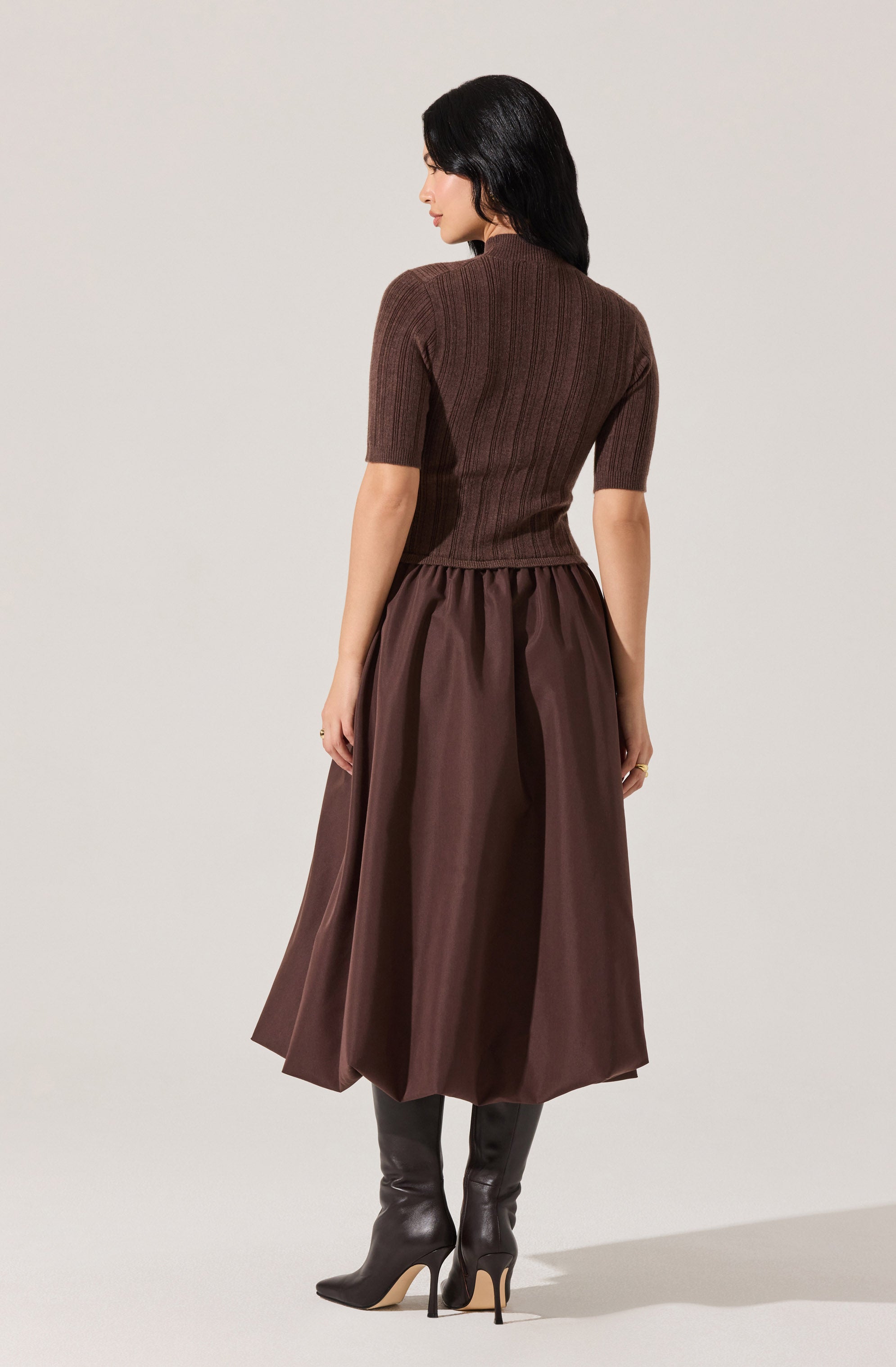 A woman is wearing a fitted, short-sleeved, ribbed knit top in a dark brown color. She pairs it with a high-waisted, full-length, dark brown silk or satin skirt that has a gathered waist. She completes the outfit with tall, black leather stiletto boots.