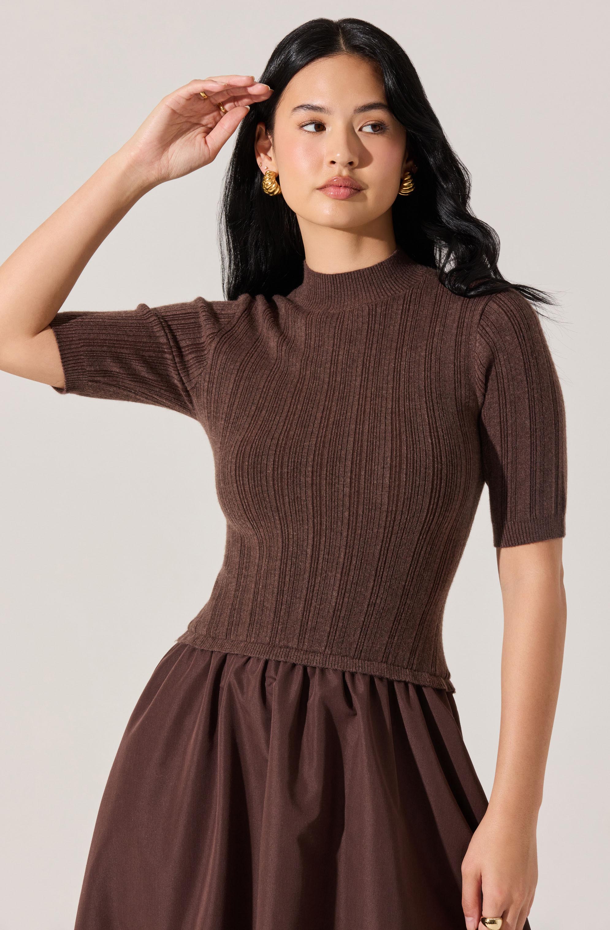 A woman is wearing a fitted, ribbed, short-sleeve knit top in a rich brown color, with a high neckline. She pairs the top with a full, gathered skirt in a matching brown shade, made of a shiny, smooth fabric. She is accessorized with large, gold, textured earrings.