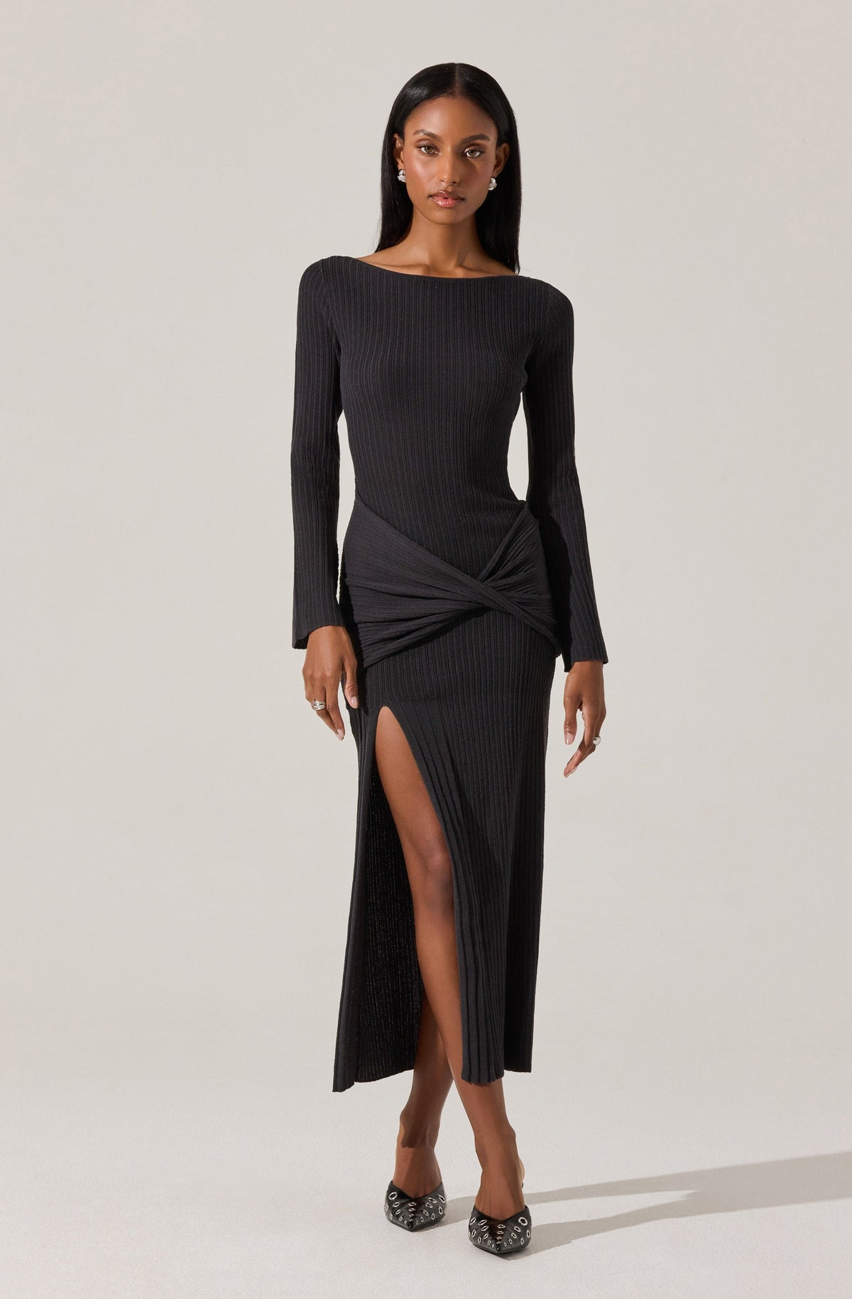 A woman is wearing a long, black, ribbed dress with a high slit on one side revealing her leg. The dress has a gathered and knotted detail at the waist, creating a draped effect. The neckline is modest and round, and the long sleeves fit snugly.