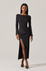 A woman is wearing a long, black, ribbed dress with a high slit on one side revealing her leg. The dress has a gathered and knotted detail at the waist, creating a draped effect. The neckline is modest and round, and the long sleeves fit snugly.