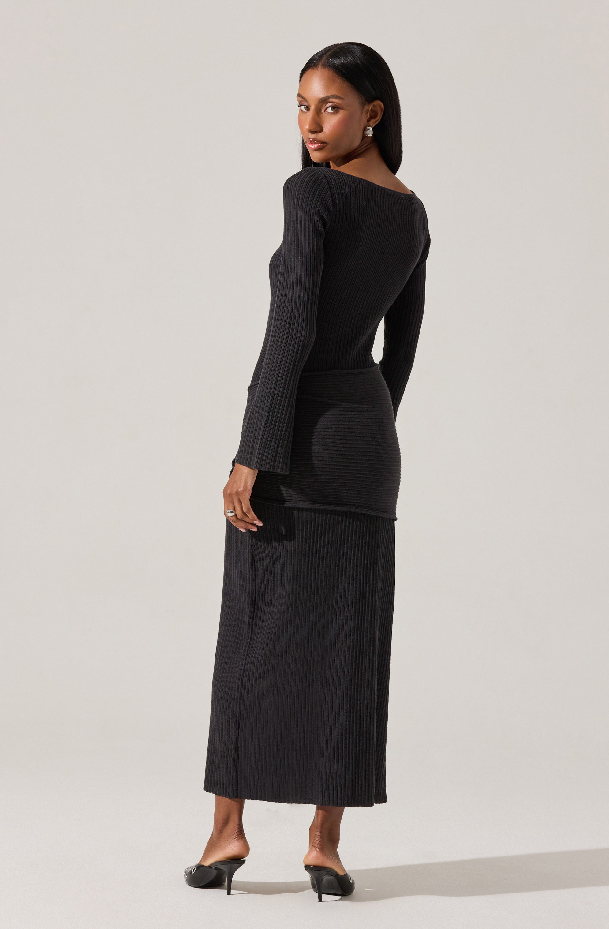 A woman is wearing a long, black, ribbed-knit dress with a fitted silhouette. The dress has a high neckline and long sleeves. She is also wearing black high-heeled shoes and earrings.
