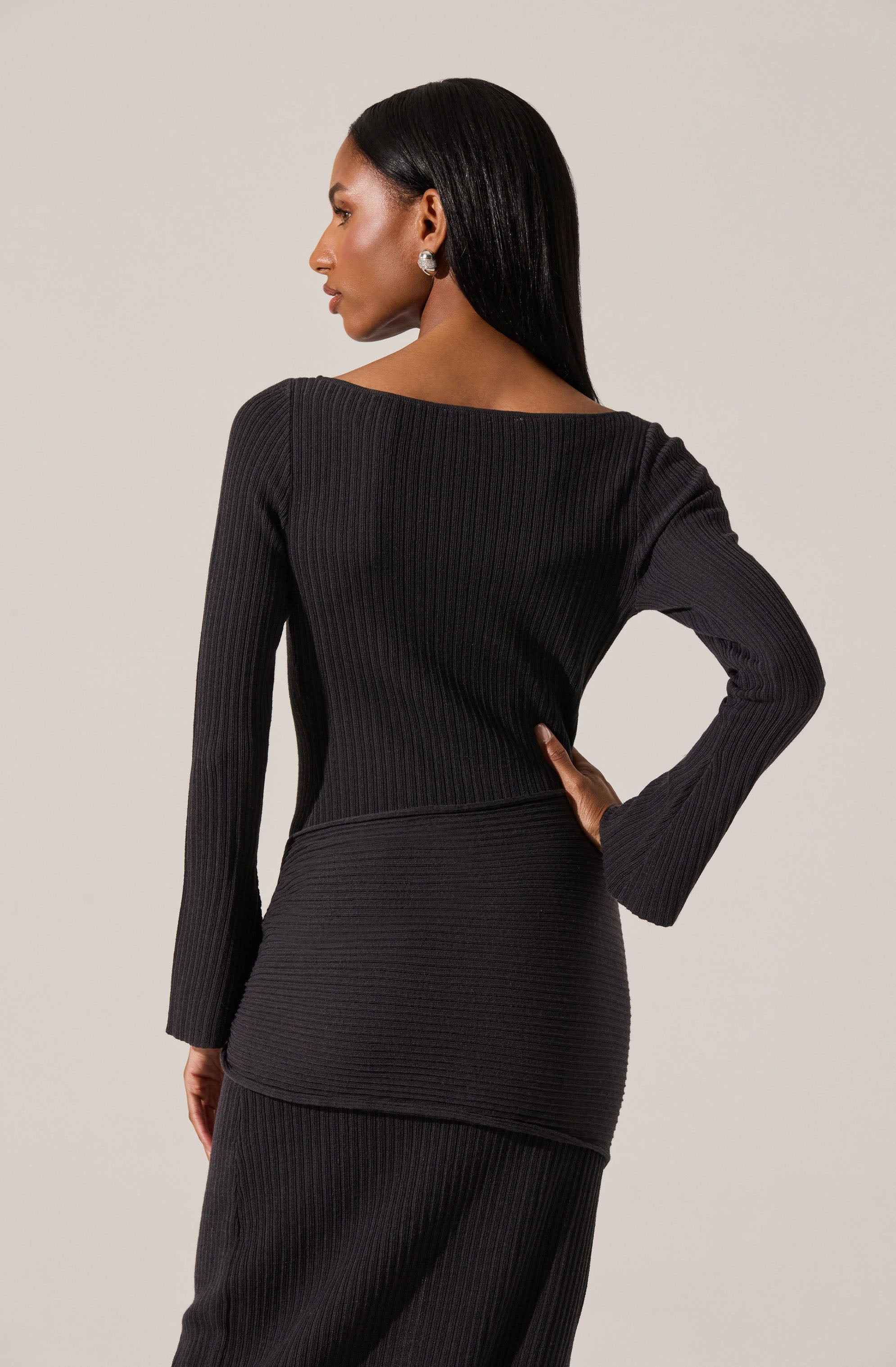A woman is wearing a black, long-sleeved, ribbed knit top and matching ribbed knit skirt. The top has a wide neckline and the outfit appears fitted, emphasizing a sleek silhouette. She accessorizes with large, round earrings and has long, straight black hair.