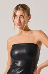 A woman is wearing a strapless black leather dress that fits snugly around her torso. The dress has a shiny, smooth texture with visible stitching lines. She accessorizes with large, circular earrings.