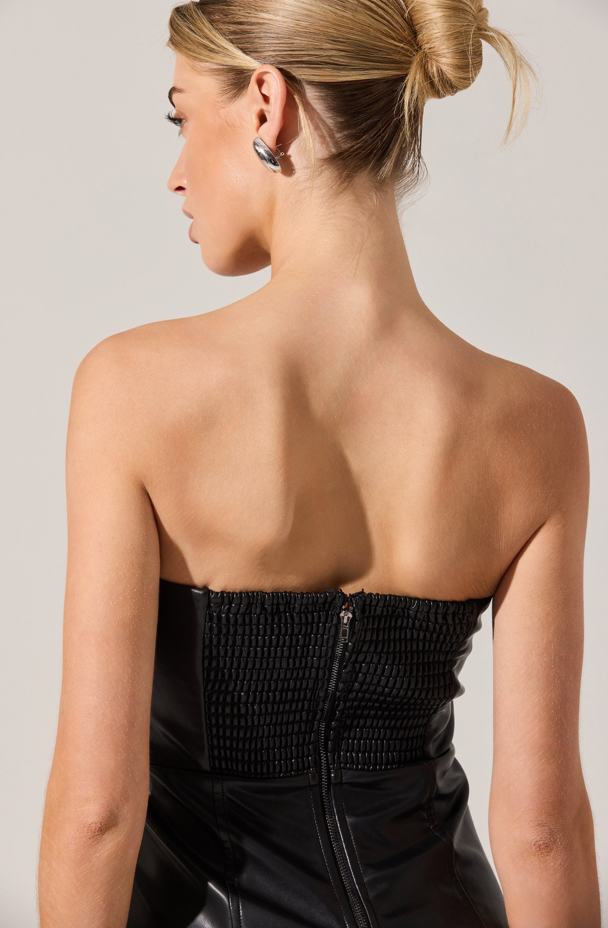 A woman with her hair in a bun is wearing a strapless black top with a textured, smocked front and a visible zipper at the back.