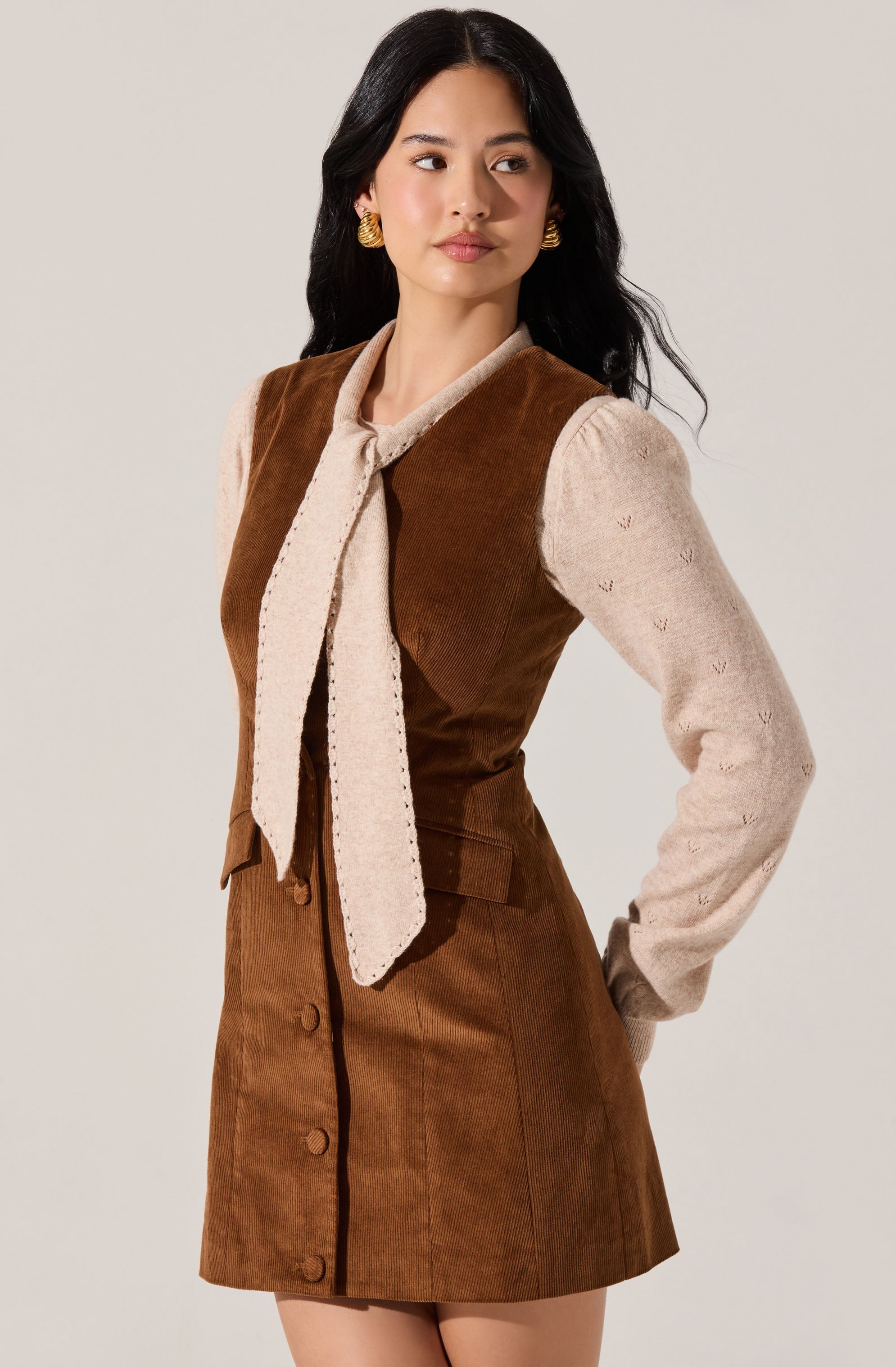 A woman is wearing a brown sleeveless dress with buttons down the front, made of a corduroy-like fabric. She has on a long-sleeved beige sweater underneath, which features small embroidered details. She accessorizes with large gold earrings and has a light beige scarf tied around her neck.