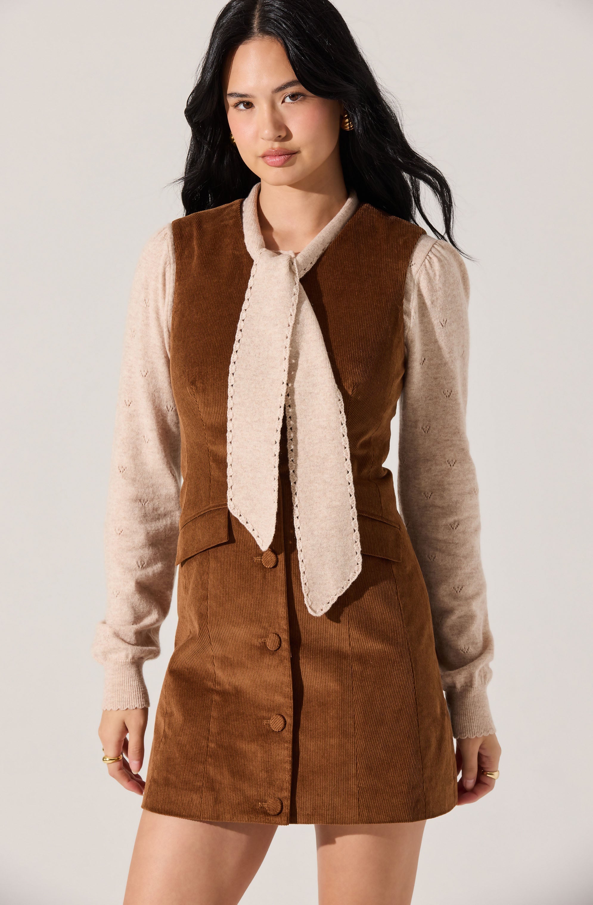 A woman is wearing a brown corduroy sleeveless dress with large buttons down the front. Underneath, she has on a beige long-sleeve sweater featuring a subtle heart pattern. She also has a matching beige scarf tied loosely around her neck.
