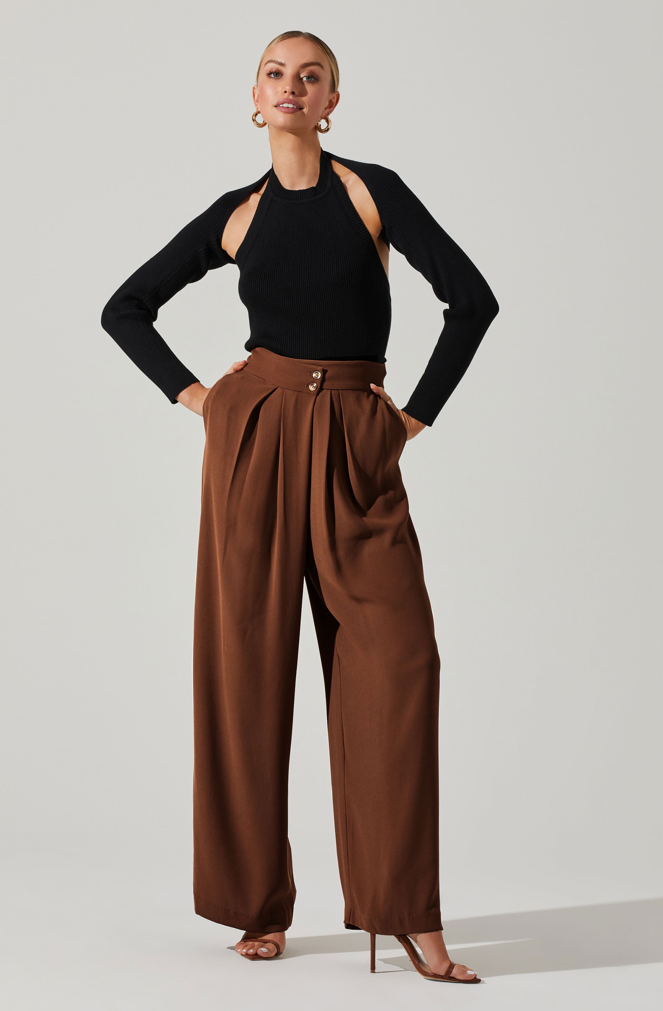 A woman wearing a black ribbed sweater with cut-out shoulders and long sleeves, paired with high-waisted brown wide-leg trousers with pleats and gold buttons at the waist. She is also wearing metallic high-heeled sandals.