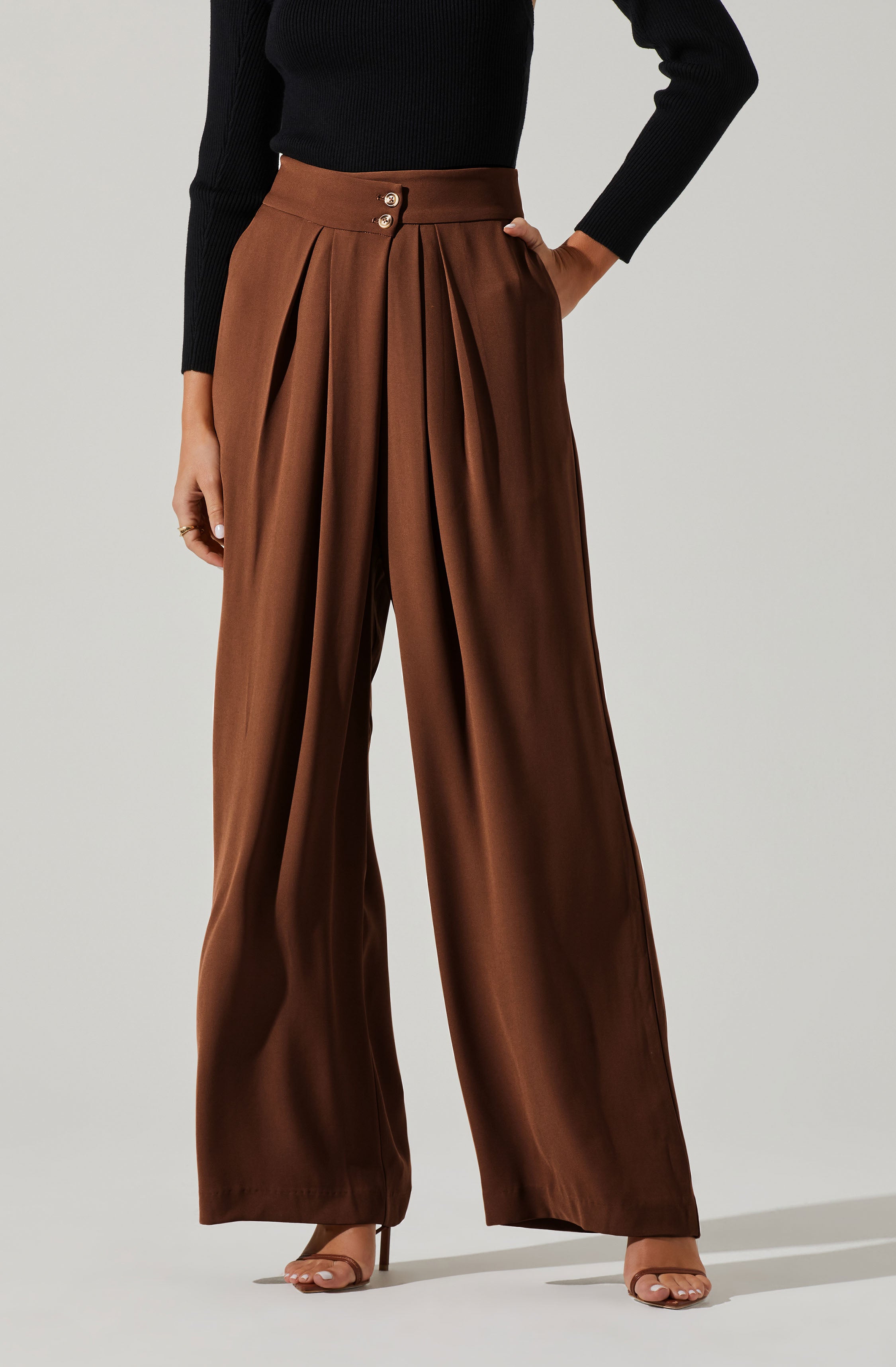 The person is wearing high-waisted, wide-leg brown satin pants with pleats at the front and a double button closure. They are also wearing a fitted black long-sleeve top.
