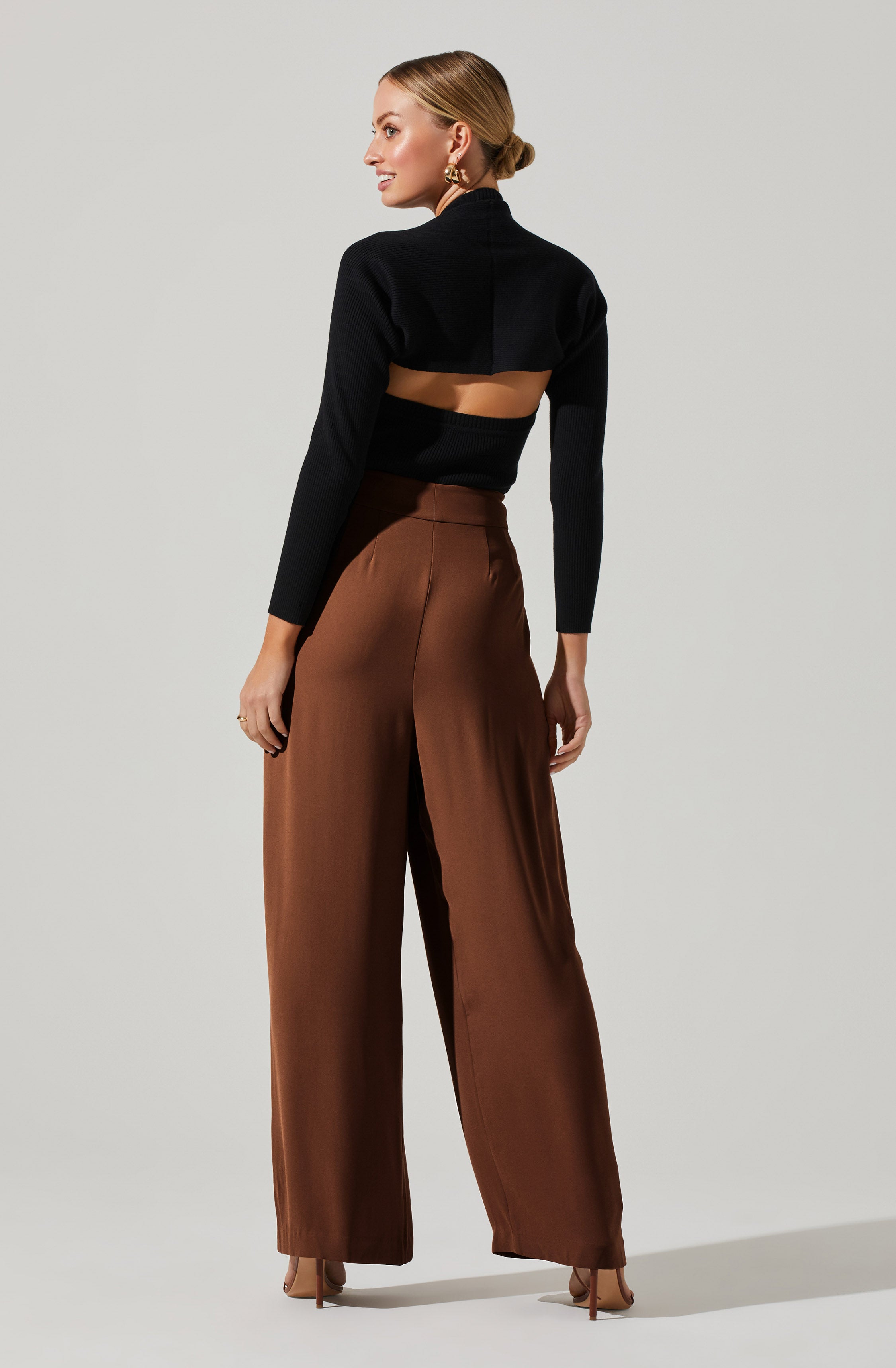 A woman is wearing a fitted black ribbed crop top with long sleeves and a high neck. The top features a cutout design at the back, exposing her mid-back. She is also wearing high-waisted, wide-leg brown satin trousers that flow down to her shoes. She has her hair styled in a low bun and is accessorized with gold hoop earrings.