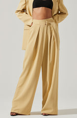 The image shows a person wearing high-waisted, wide-leg beige trousers with pleats at the waist and decorative buttons. The person is also wearing a black crop top and a matching beige blazer. The outfit appears stylish and formal.