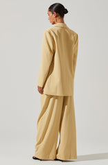 The woman is wearing a beige suit consisting of a long blazer with a notched collar and wide-leg trousers. The blazer has a structured fit with shoulder pads, and the trousers are high-waisted and flowing, creating a relaxed silhouette. She accessorizes with large gold hoop earrings.