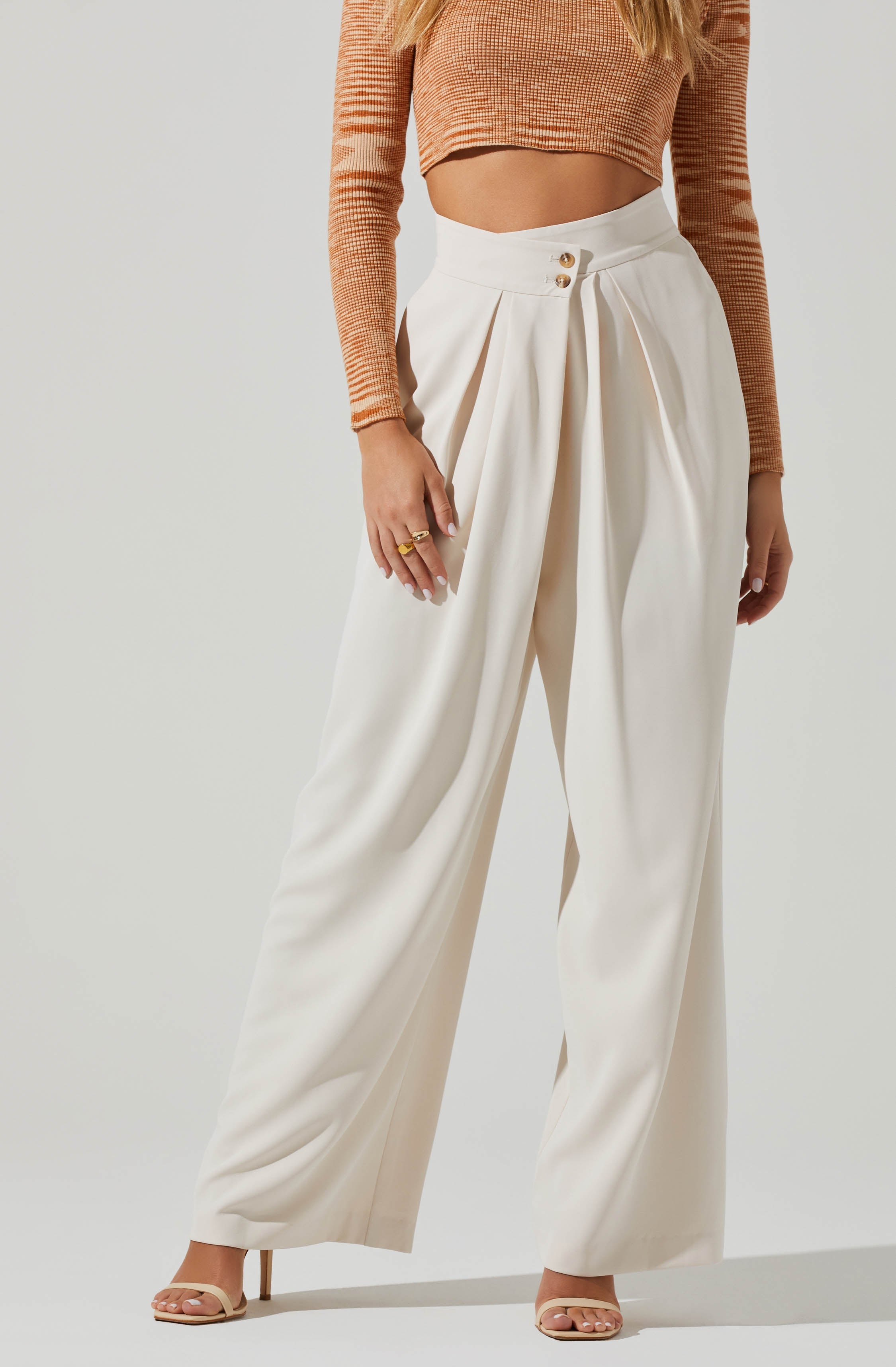 The image shows a woman wearing high-waisted, wide-leg white trousers with pleats, paired with a long-sleeved, fitted crop top in a light orange and beige striped pattern. She has nude high-heeled sandals on her feet and is accessorizing with gold rings on her fingers.