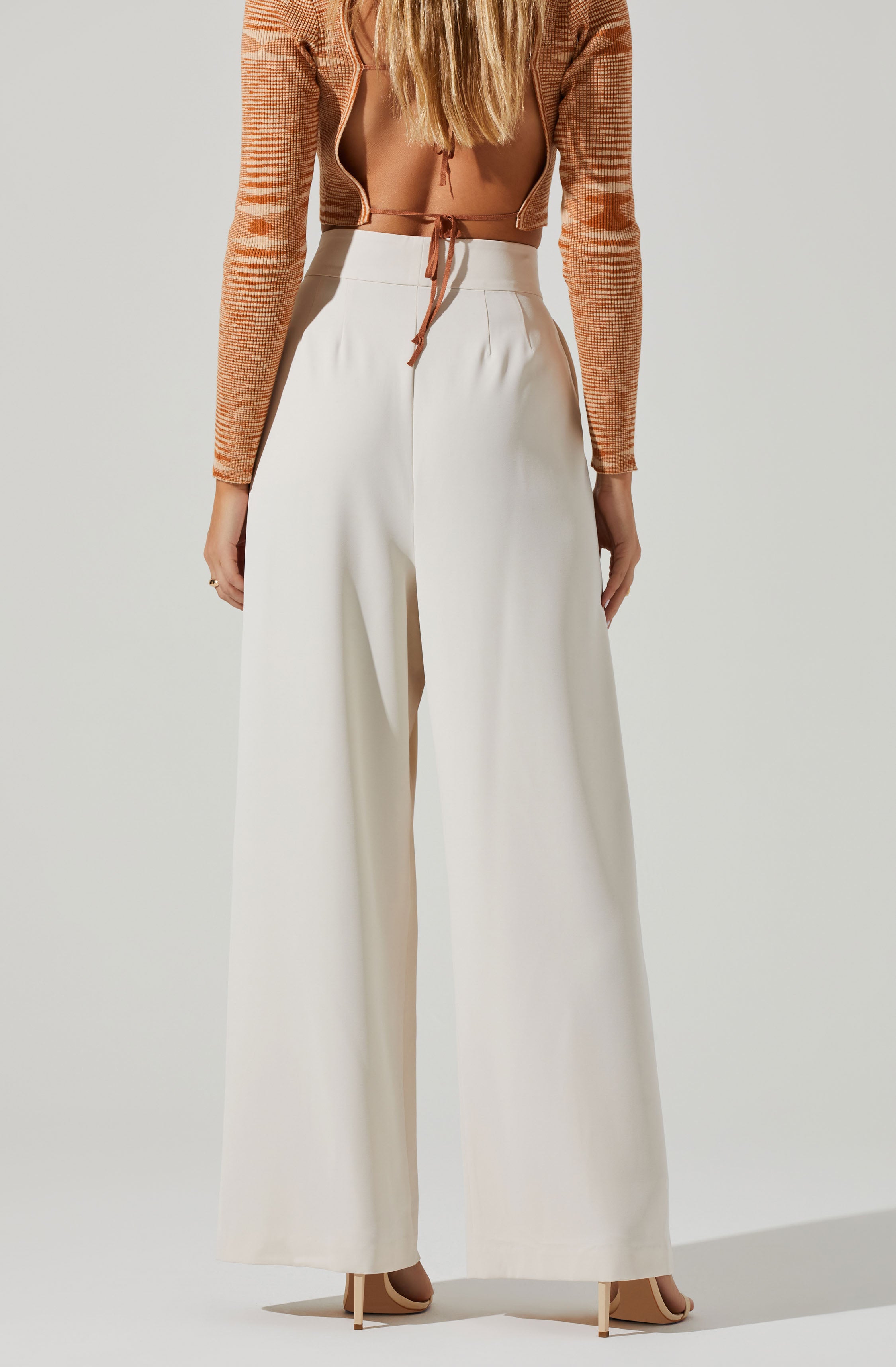 The image shows a person dressed in high-waisted, wide-leg white pants that reach the ground, paired with nude high heels. They are wearing an orange and beige patterned, long-sleeved, cropped cardigan with a zipper in the front, which is open, revealing a matching orange bralette underneath. The cardigan has a small tie at the bottom center.