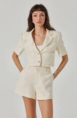 A woman is wearing a cream-colored, textured two-piece outfit consisting of a cropped blazer with short sleeves, large front pockets, and two decorative buttons, paired with matching high-waisted shorts. The blazer has a notched lapel collar and the fabric appears to have a woven or boucle texture. The outfit exudes a chic, sophisticated style.