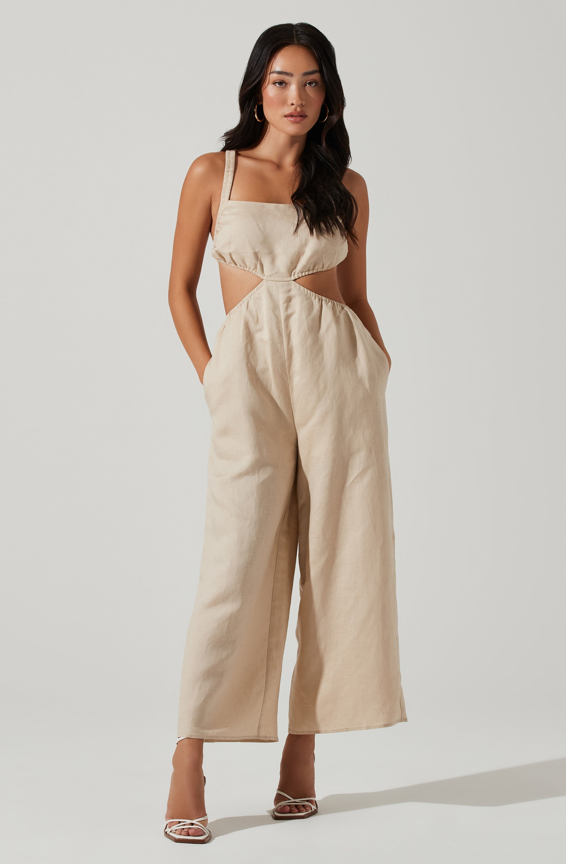 A woman is wearing a beige, sleeveless jumpsuit with cut-out details on the sides at the waist and a square neckline. The fabric appears to be lightweight and slightly textured. She has paired the outfit with open-toe high heels and gold hoop earrings.