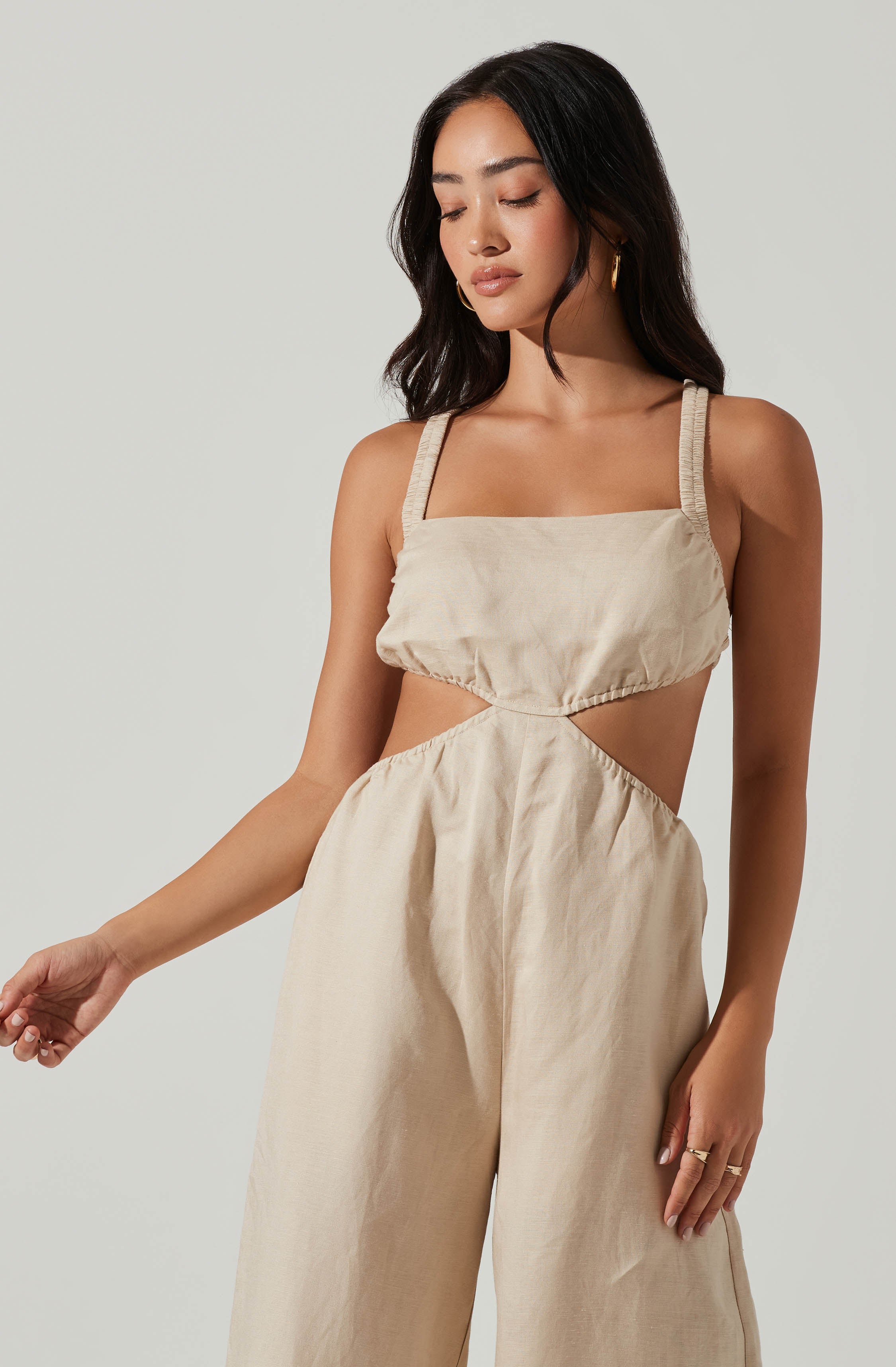 A woman is wearing a beige, sleeveless jumpsuit with wide-leg pants. The top part features a square neckline and wide, gathered straps, with a cutout design that shows her midsection. The fabric appears lightweight with a slightly textured finish. She has long, dark hair styled in loose waves and is accessorizing with gold hoop earrings and rings on her fingers.