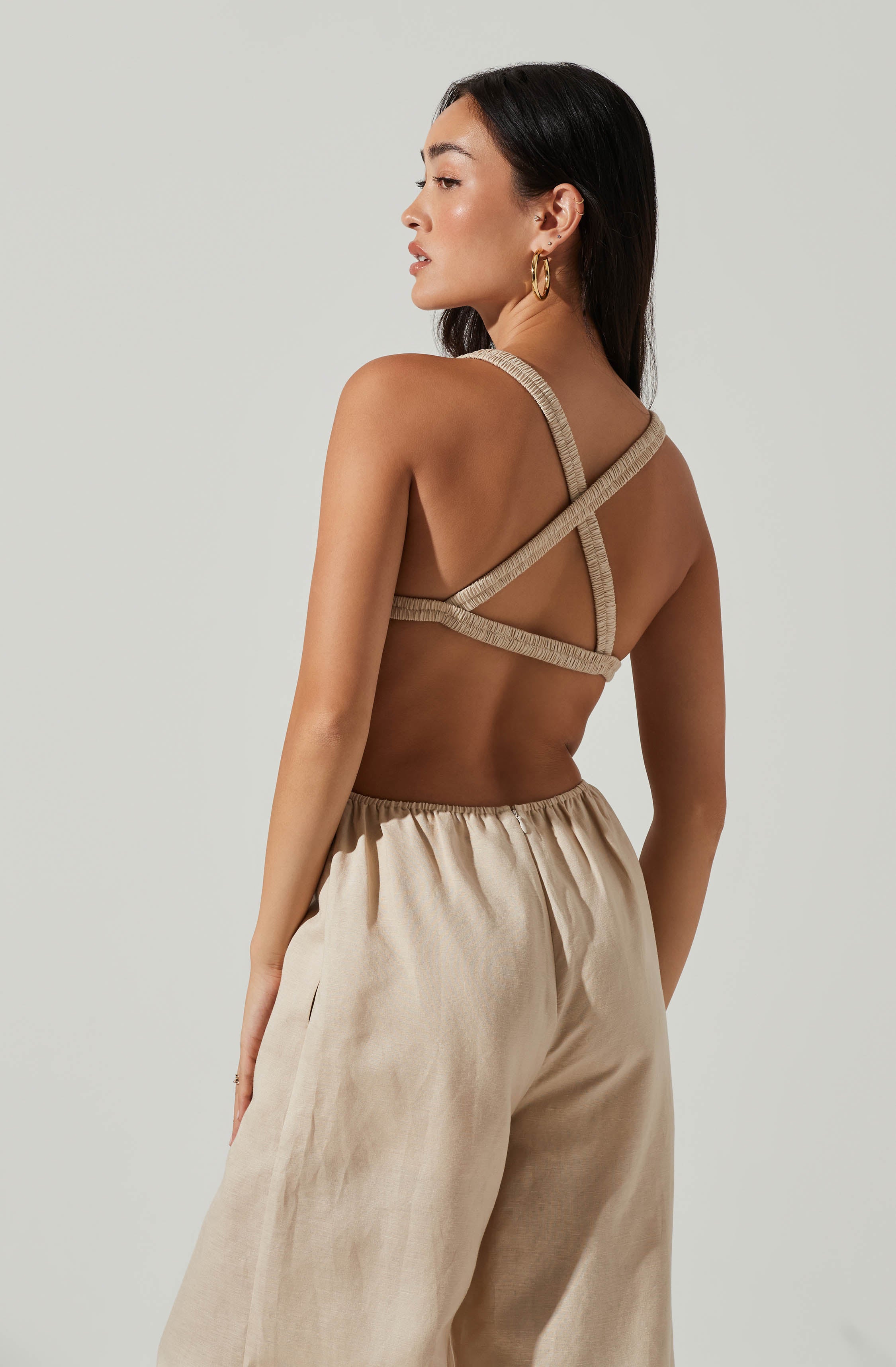 The woman is wearing a beige, textured, crisscross halter top with multiple straps around her shoulders and back. She pairs it with high-waisted, loose-fitting beige pants made of a lightweight fabric.