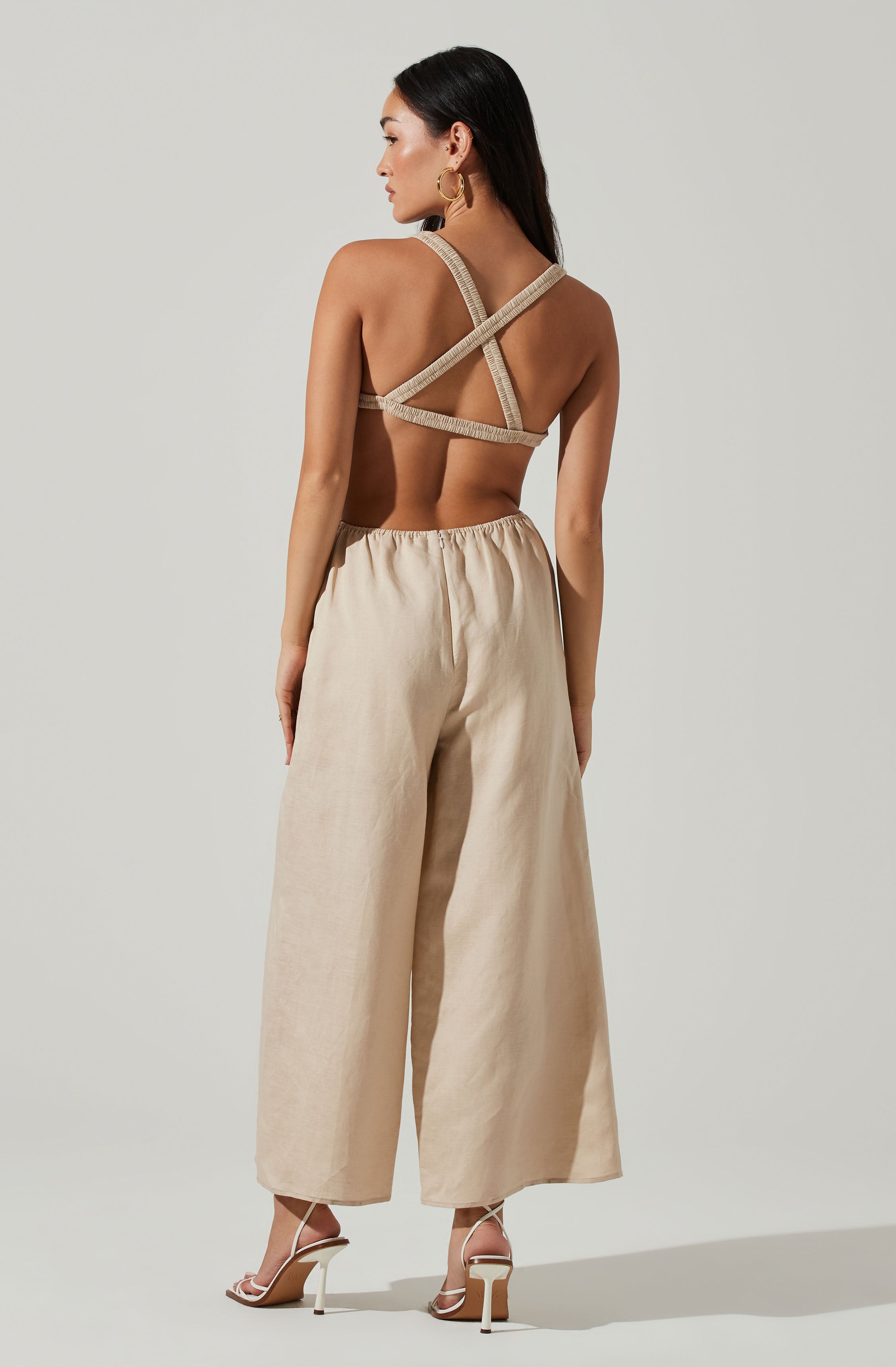 A woman is wearing a beige halter-style top with crisscross straps on the back and a pair of wide-leg beige linen pants. She is also wearing white high-heeled sandals and gold hoop earrings.