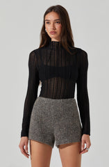 A young woman is wearing a black, long-sleeved, semi-sheer, high-neck top with vertical lace patterns. She pairs it with high-waisted, gray, textured shorts.