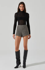 A woman is wearing a black, long-sleeved, semi-sheer, high-neck top with vertical stripes. She pairs it with gray, high-waisted, tailored shorts with a textured fabric. She completes the outfit with knee-high black leather boots with pointed toes and stiletto heels.