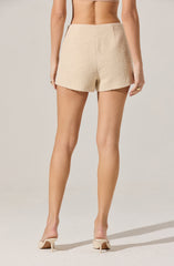 A person wearing high-waisted, textured, cream-colored shorts with a subtle shimmer, paired with beige high-heeled shoes. The shorts have a slightly frayed hem and are made of a boucle or tweed-like fabric.