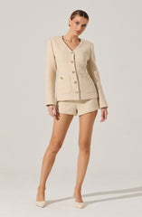 A woman is wearing a cream-colored tweed suit consisting of a fitted blazer with gold buttons and two front pockets, paired with matching high-waisted shorts. She is also wearing beige pointed-toe high heels.