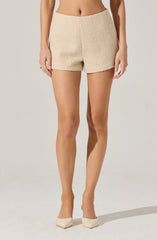 A woman is wearing beige textured shorts that have a slightly frayed hem, paired with beige high-heeled shoes. Her outfit exposes her midriff and her arms.