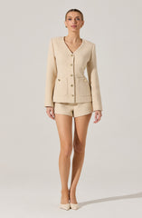 The woman is wearing a cream-colored, textured, tailored suit consisting of a fitted blazer with a V-neckline, gold buttons down the front, and two front pockets, paired with matching high-waisted shorts. She is also wearing beige high-heeled shoes.