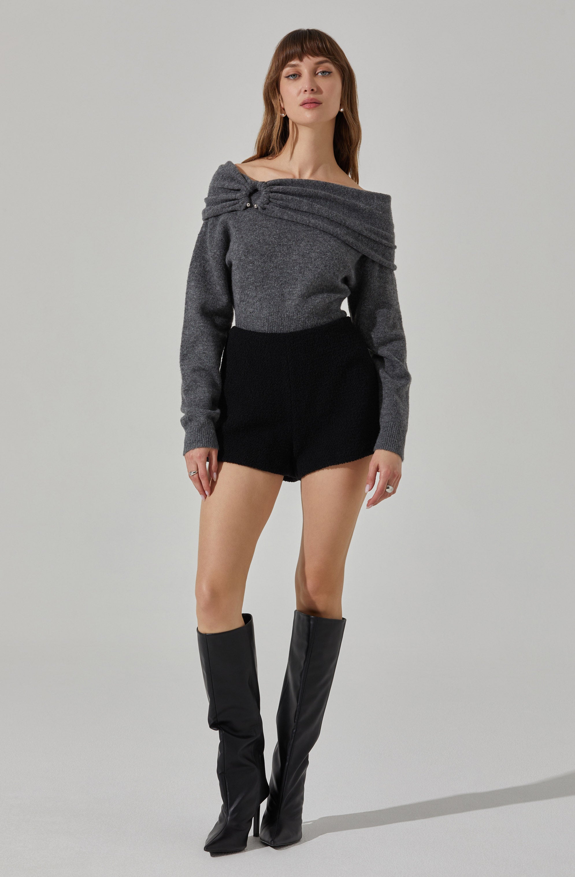 A woman is wearing a off-the-shoulder gray sweater with a wrap detail and a small decorative pin on the shoulder, paired with high-waisted black shorts and knee-high black boots.