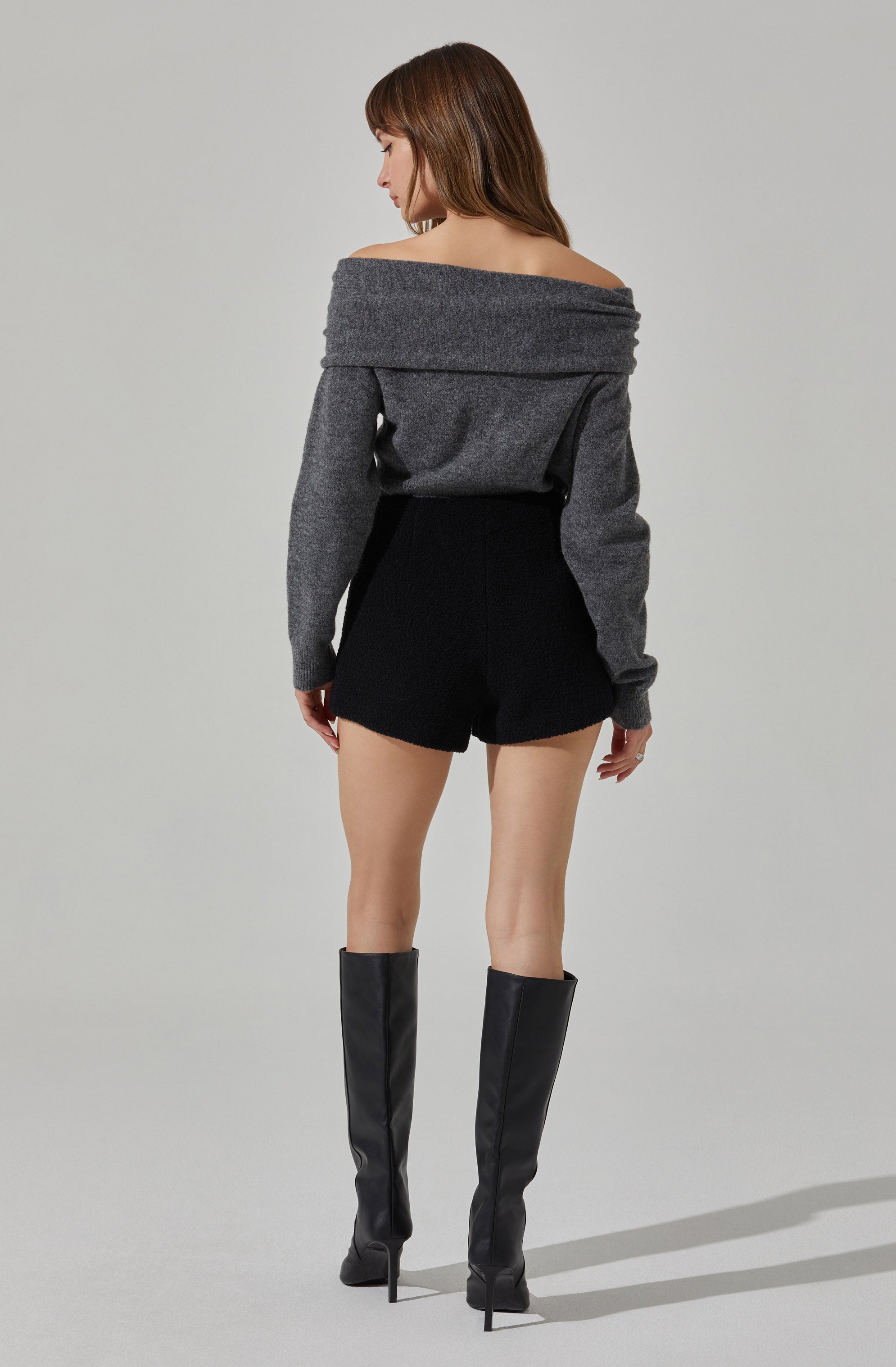 A woman is wearing an off-the-shoulder, long-sleeved, gray sweater paired with high-waisted black shorts. She has on tall black high-heeled boots.
