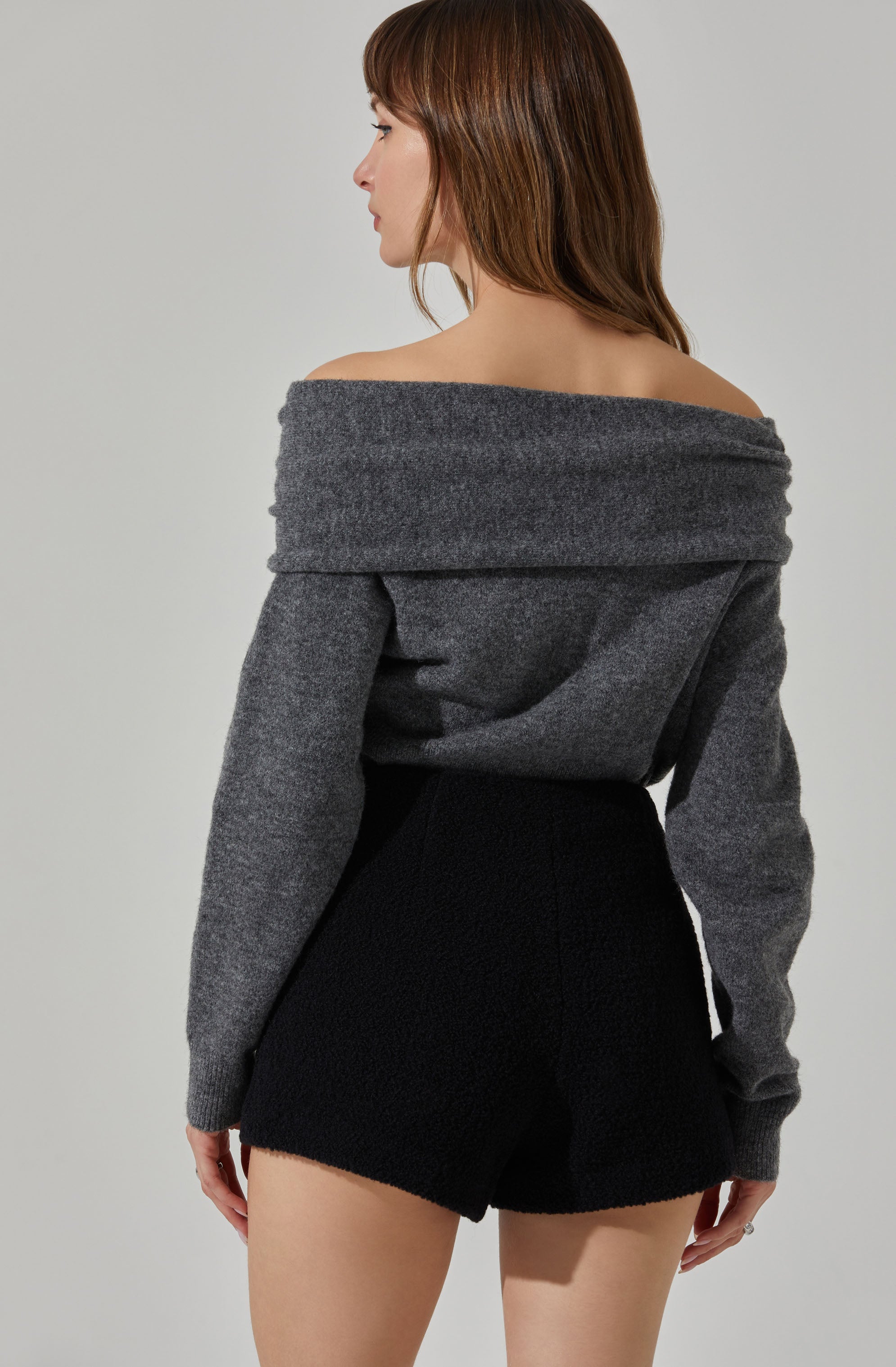 A woman is wearing an oversized, off-the-shoulder, gray sweater with a wide neckline that drapes over her shoulders. She is also wearing high-waisted, black textured shorts that fit snugly.