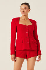 A woman is wearing a bright red textured suit consisting of a fitted blazer with a square neckline, long sleeves, and four large buttons down the front. The blazer has two flap pockets on the front. She is also wearing matching red textured shorts. The outfit has a tailored, sophisticated style.