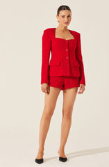 A woman is wearing a bright red, textured, tailored suit set consisting of a fitted blazer with shoulder pads, a matching crop top underneath, and high-waisted shorts. The blazer has a structured collar, long sleeves, and large buttons down the front, with two flap pockets on the front. She is also wearing black pointed-toe shoes.