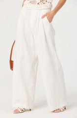 The image shows a person wearing high-waisted, wide-leg white pants with a relaxed fit. The pants have pleats at the waist and side pockets. The person is also wearing a white, sleeveless, crochet top with floral patterns. They have their right hand in their pocket and are wearing white strappy high-heeled sandals. A brown handbag with a thin strap is partially visible hanging on the person's left side.