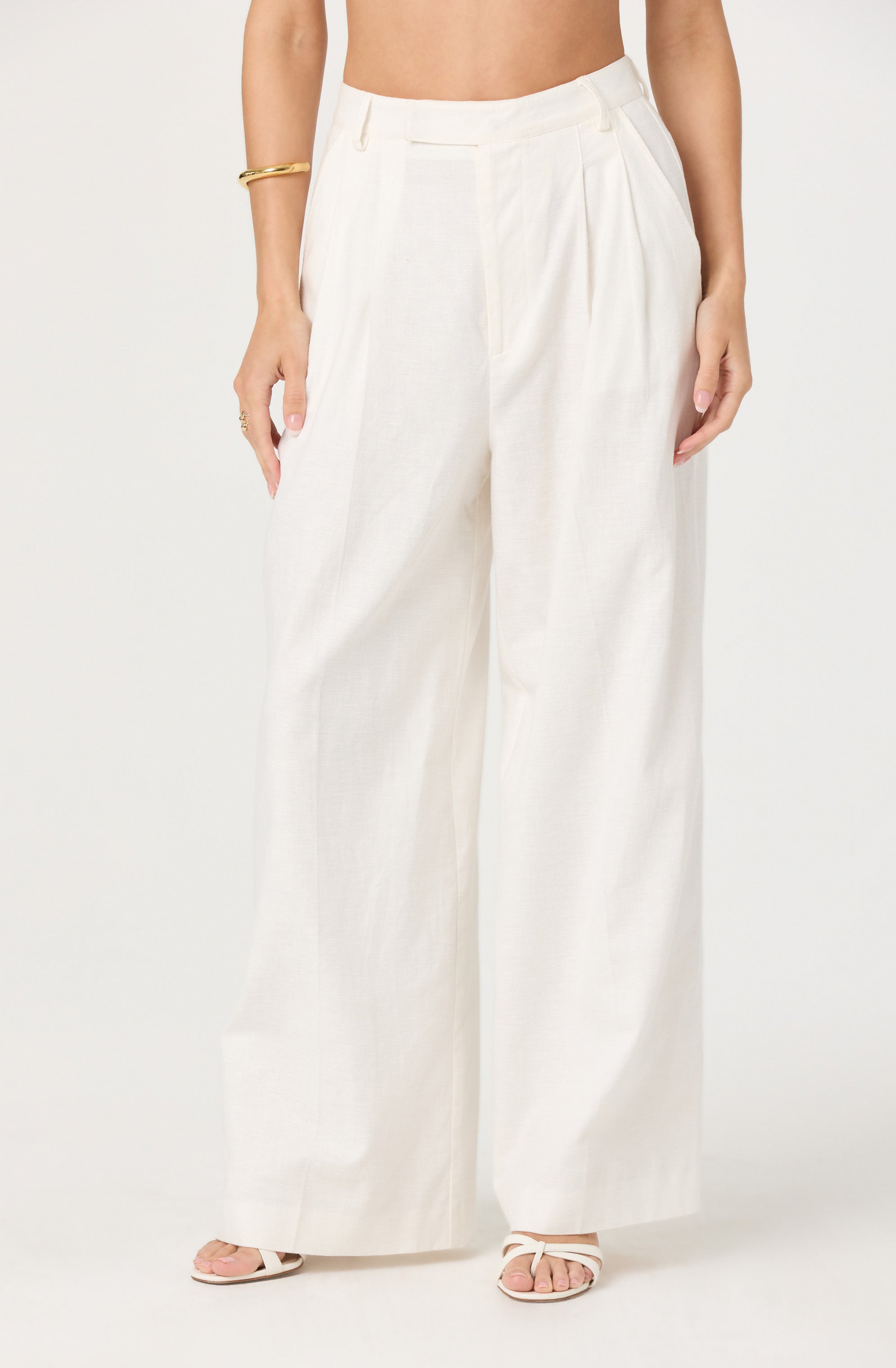 A person is wearing high-waisted, wide-leg white trousers made from a light, flowy fabric. The trousers have a tailored waistband with belt loops and pleats at the front. The person is also wearing a gold bracelet on their right arm and white strappy sandals.